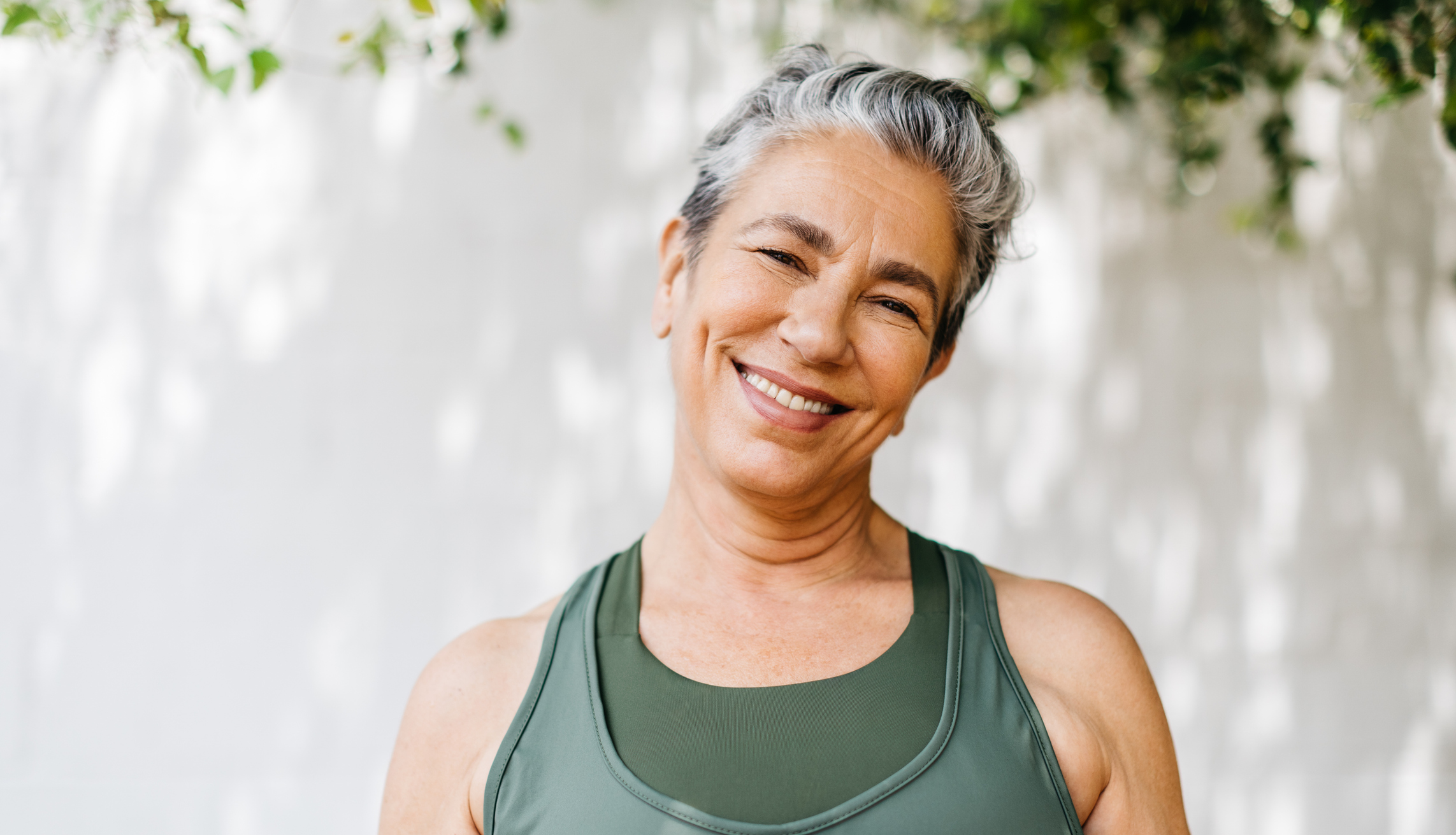 Smiling woman with short gray hair wearing a green athletic tank top, standing outdoors against a light wall with soft greenery.